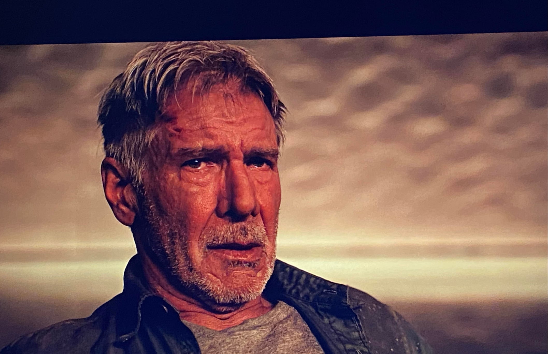 An older man with gray hair and a beard looks emotional and weary, sitting in front of a blurred, sepia-toned background. He wears a gray shirt and an open jacket.