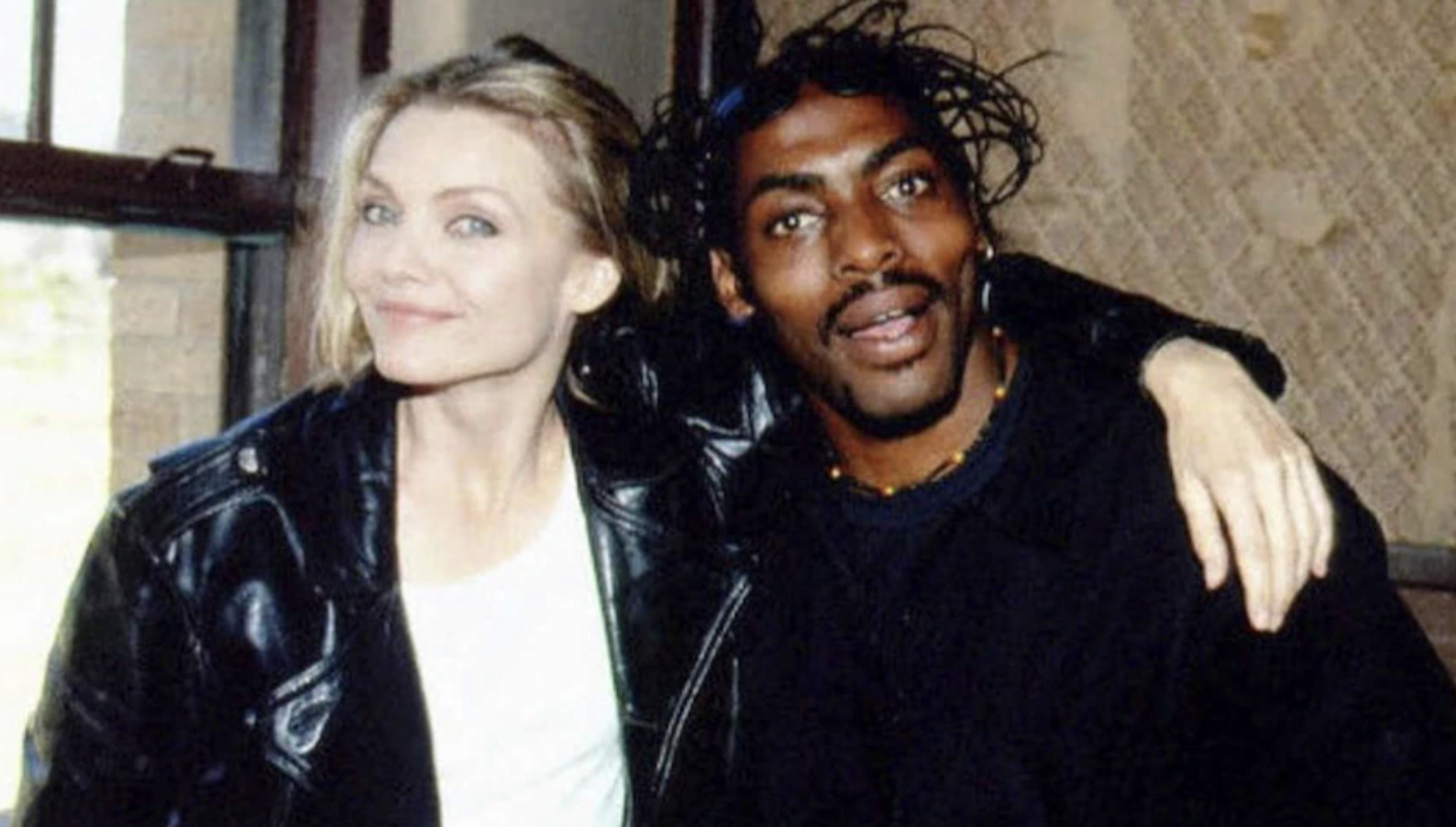 Two people pose together indoors. The person on the left has light hair, wears a black leather jacket, and smiles at the camera. The person on the right has dark hair in braids and wears a black shirt, with one arm around the other.