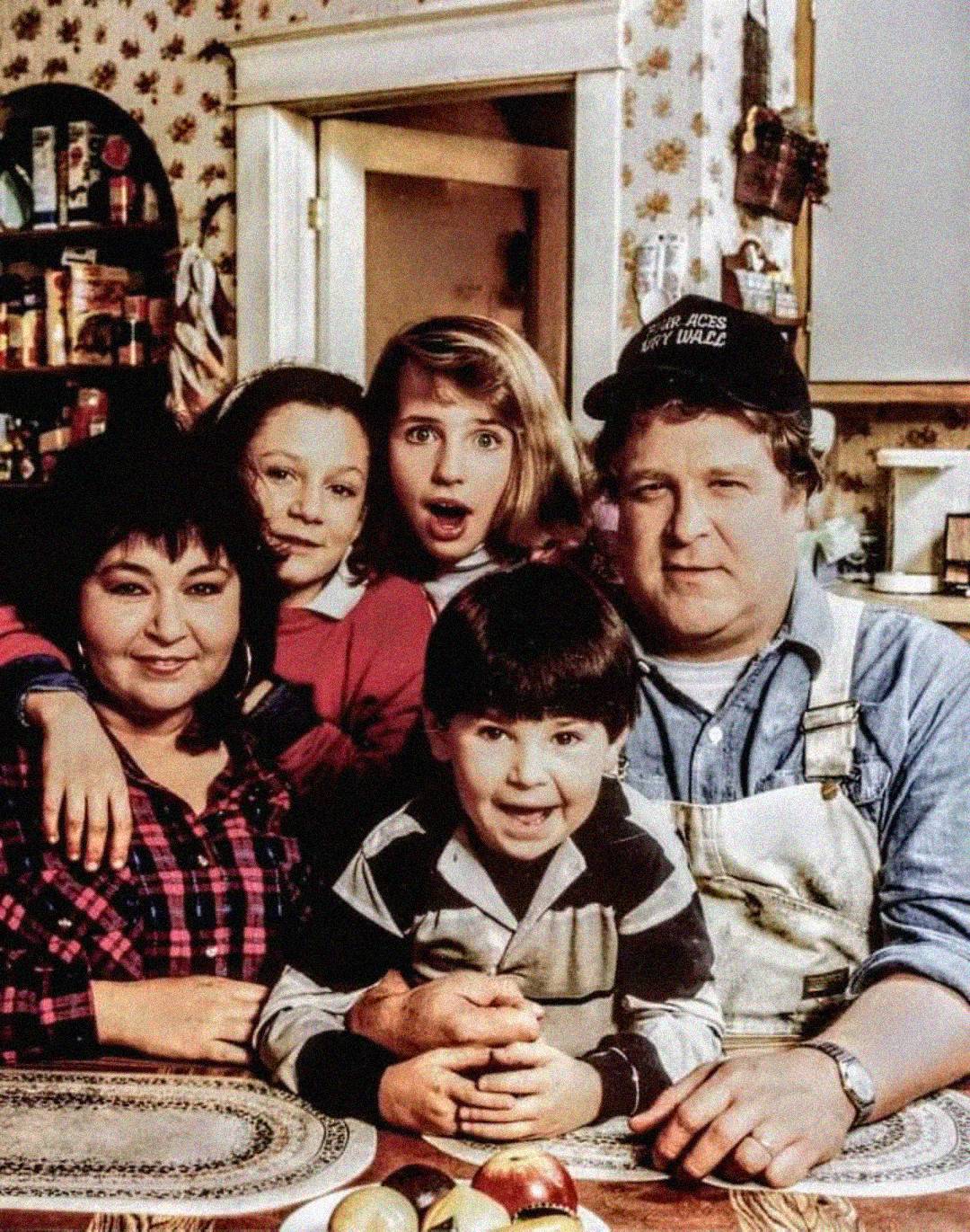 A family of five poses in a cozy kitchen. Three adults and two children smile and lean close together, with shelves of food and floral wallpaper in the background. The scene feels warm and nostalgic.