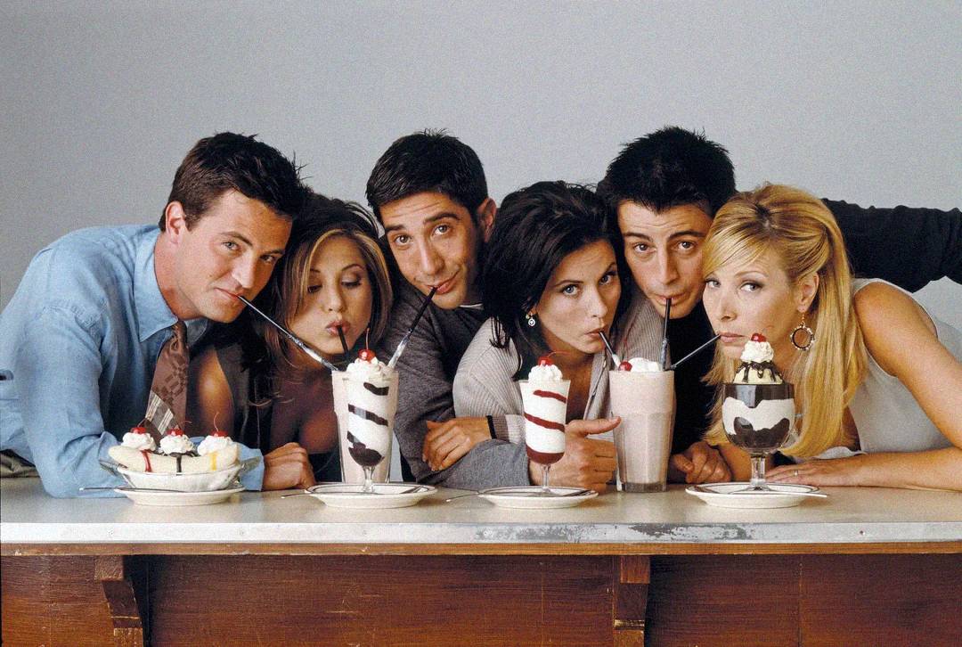 Six people sit closely together at a counter, each drinking milkshakes or eating desserts. They lean in and smile while sharing straws and desserts in a friendly, lighthearted atmosphere.