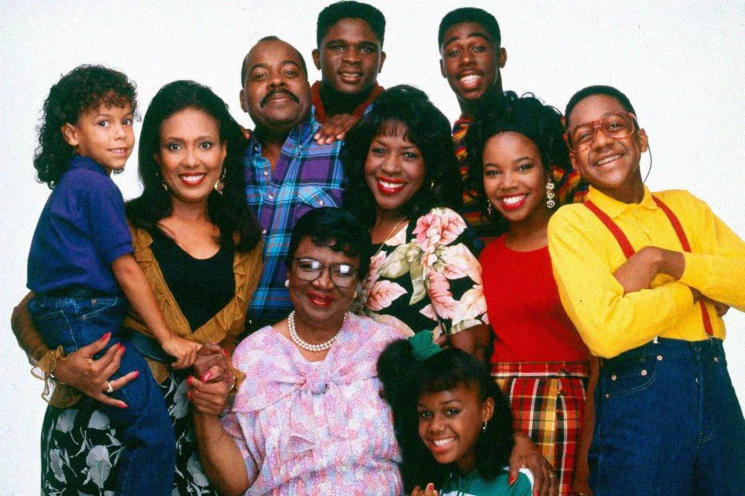 A group of ten people, including adults, teenagers, and one child, pose together and smile against a plain white background. They wear colorful, casual 90s clothing and appear happy and close-knit.