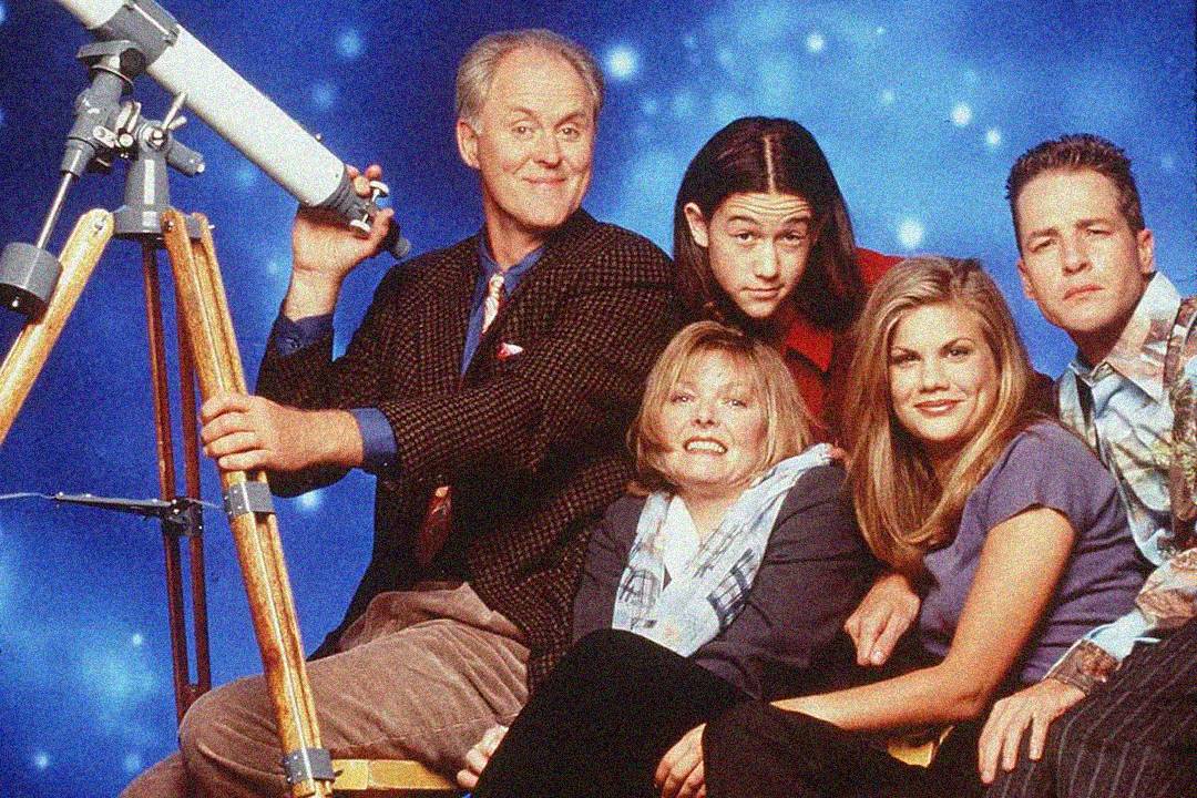 Five people pose together in front of a blue, starry background. One person holds a telescope. The group is smiling and sitting closely, dressed in casual 1990s-style clothing.