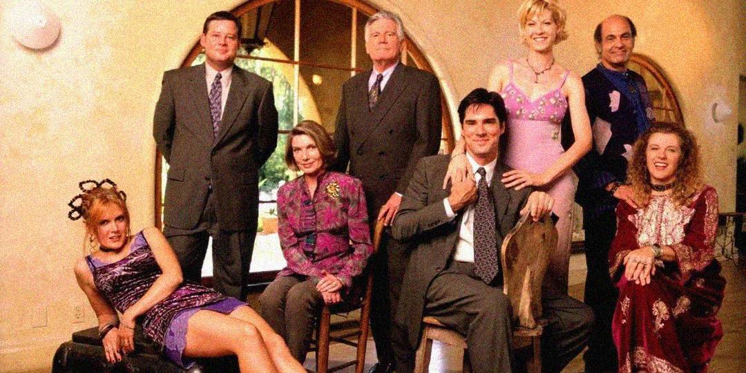Eight people dressed in colorful, formal 1990s attire pose indoors, some sitting and some standing, in front of a large arched window with a bright outdoor view. The mood is casual and cheerful.