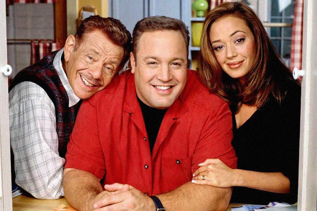 Three people are smiling together indoors. The man in front wears a red shirt, while the man on the left wears a vest and the woman on the right has long hair and is gently resting her hand on the man's arm.