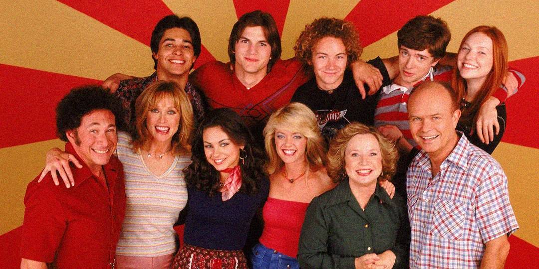 A group of eleven people, smiling and posing together in front of a red and yellow radial background. Some have their arms around each other, and the group includes both men and women of varying ages.
