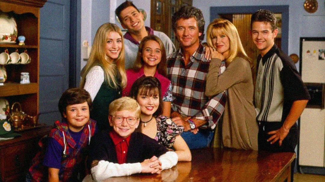 A group of eight people, including adults and children, pose together smiling around a wooden table in a cozy, homey kitchen setting.
