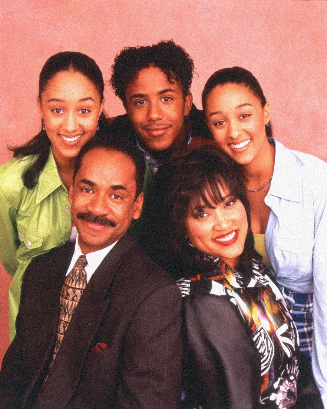 Five people pose closely together and smile at the camera against a pink background; three youths stand behind two seated adults, all dressed in colorful, late 1990s-style clothing.