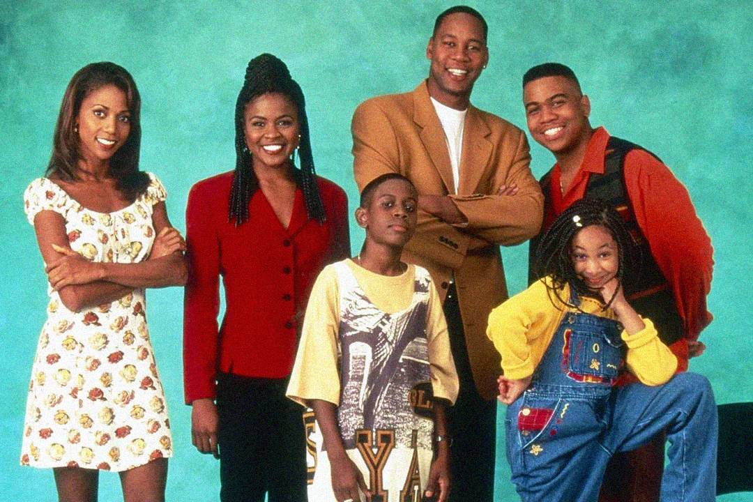 Six people posing in front of a teal background. Four adults stand behind, smiling, while two kids stand in front; the girl on the right is playfully posing in blue overalls and a yellow shirt.