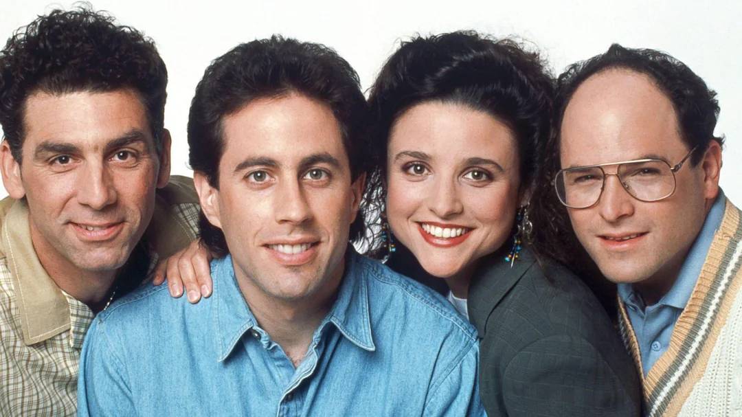Four people pose closely together, smiling at the camera. The group includes three men and one woman, all dressed in casual 1990s-style clothing, against a plain white background.