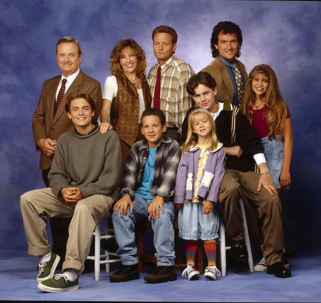 A group of eight people, including adults and children, pose together in two rows against a blue studio backdrop. Some are sitting on stools while others stand behind them, all smiling at the camera.