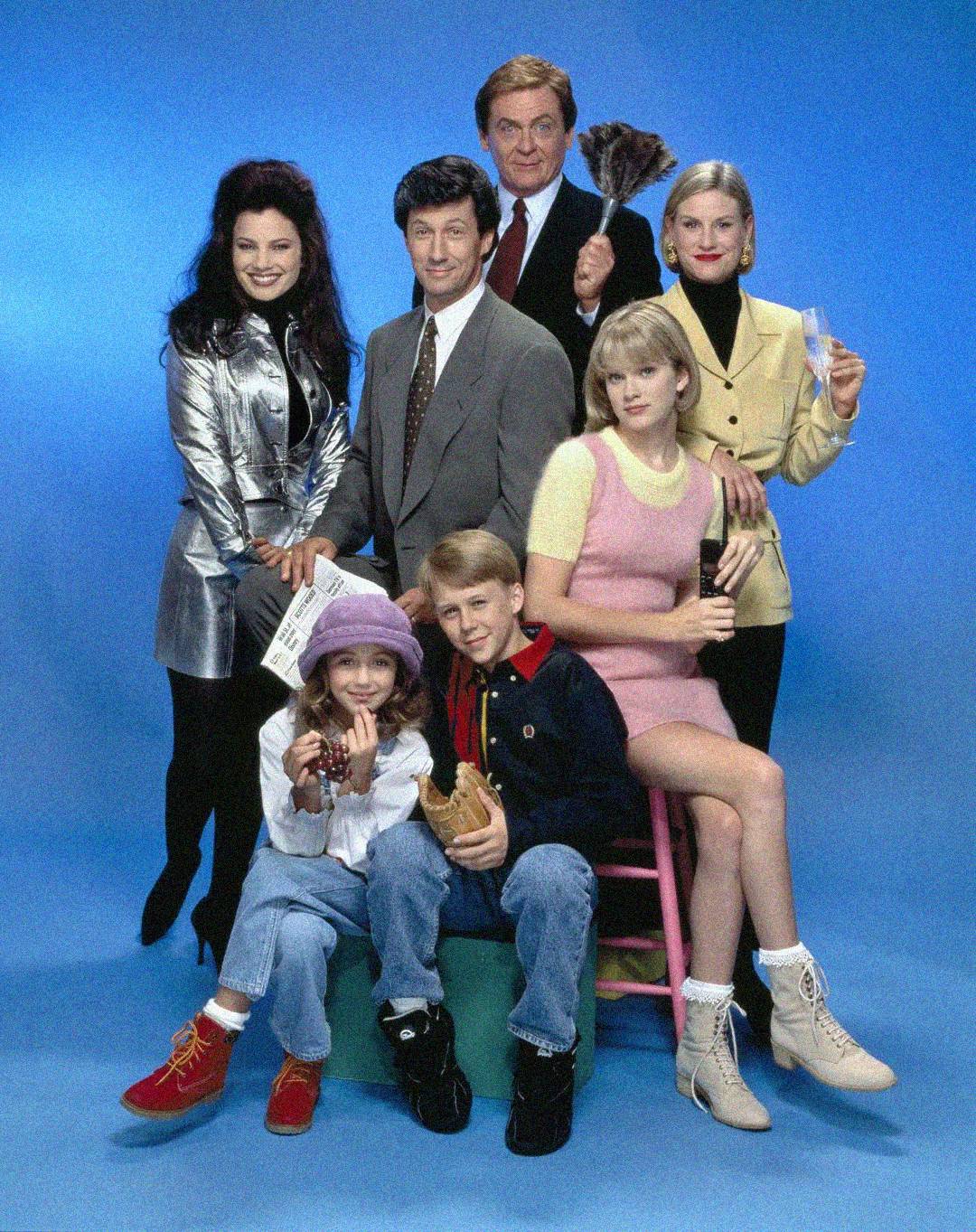 A group of six adults and one child pose together against a blue background, dressed in colorful 1990s clothing. Some are standing, some are seated, and they are smiling and looking at the camera.