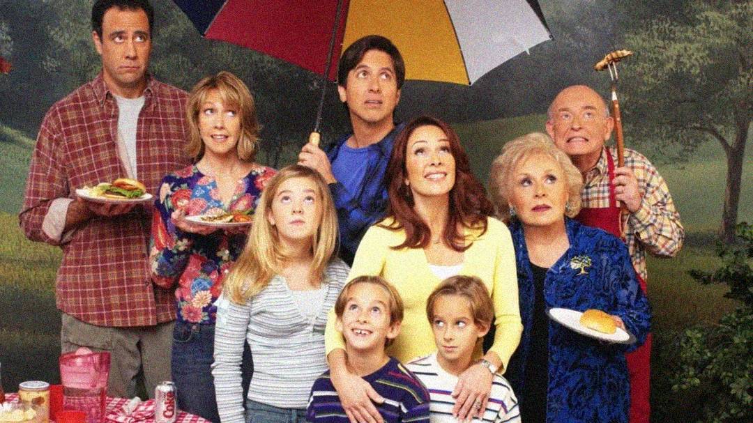 A family of eight people poses outdoors for a photo. Some smile while others look surprised. One man holds an umbrella, another grills food, and plates with burgers are visible, suggesting a picnic or barbecue.