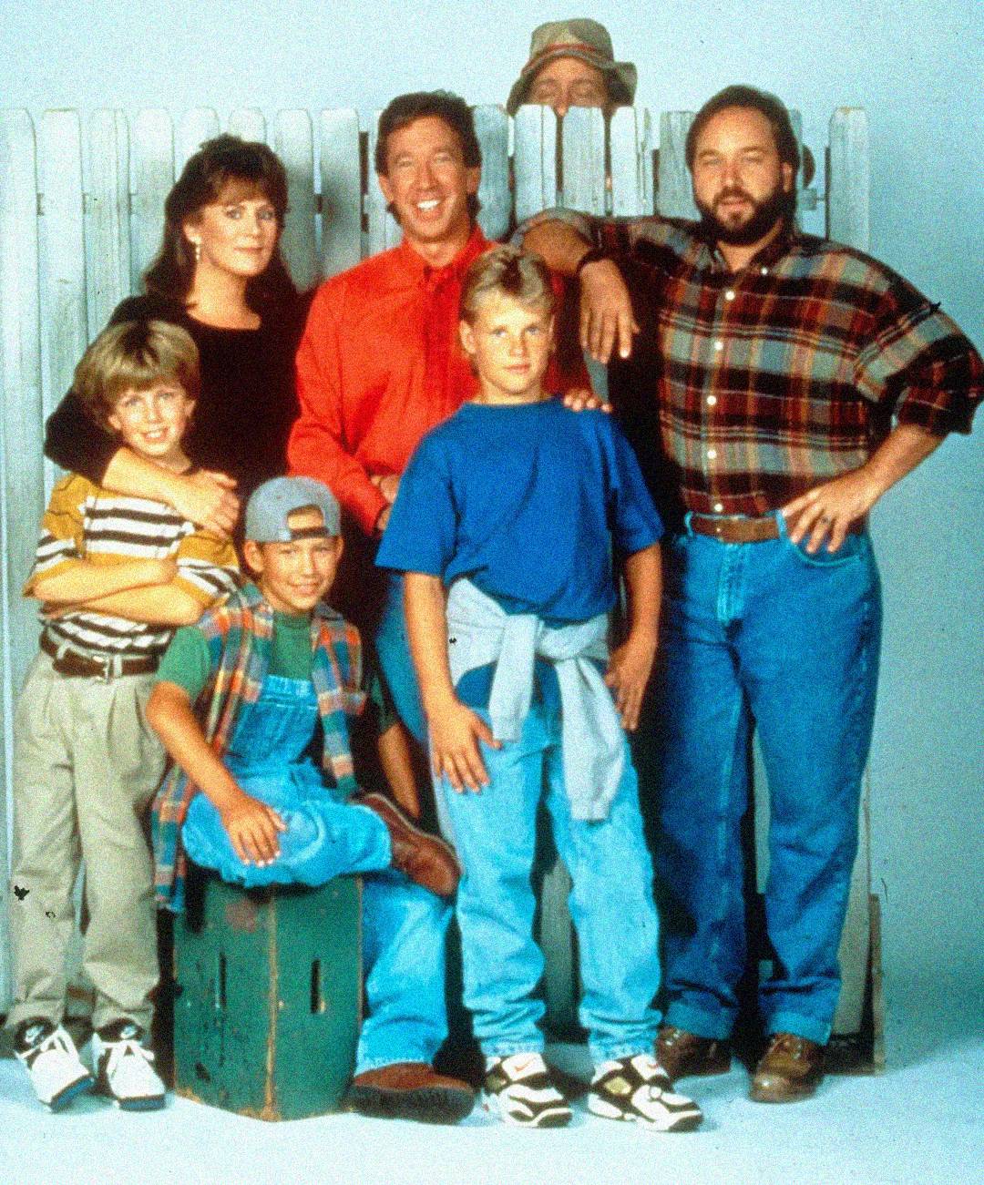 A group of seven people, including adults and children, stand and sit in front of a white picket fence, smiling at the camera. One person peeks over the fence in the background.