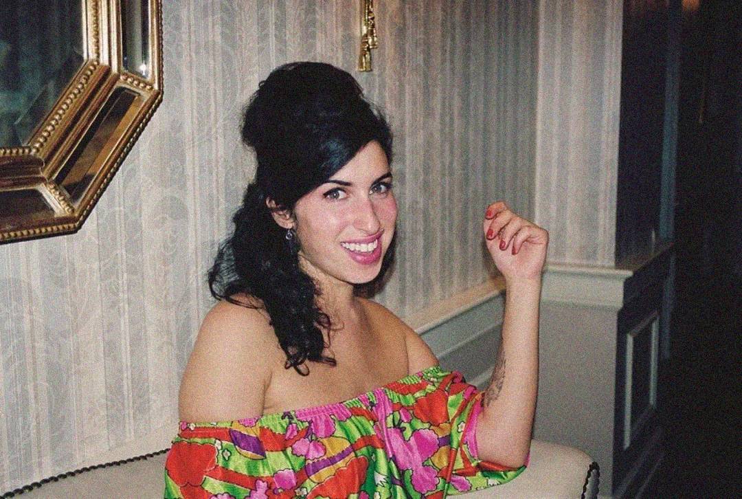 A woman with dark hair in a beehive style, wearing a colorful off-shoulder floral dress, sits on a white couch and smiles at the camera in a vintage-style room with patterned wallpaper and a gold-framed mirror.