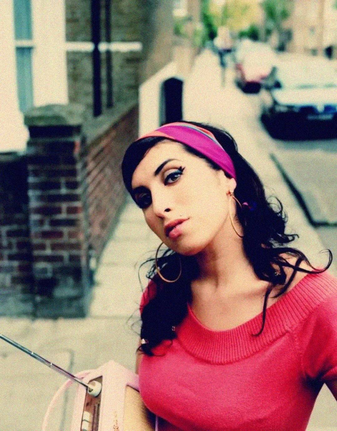 A woman with dark hair, wearing a pink headband and off-the-shoulder top, stands on a city sidewalk holding a retro radio. She looks at the camera with a slight tilt of her head; the background is slightly blurred.