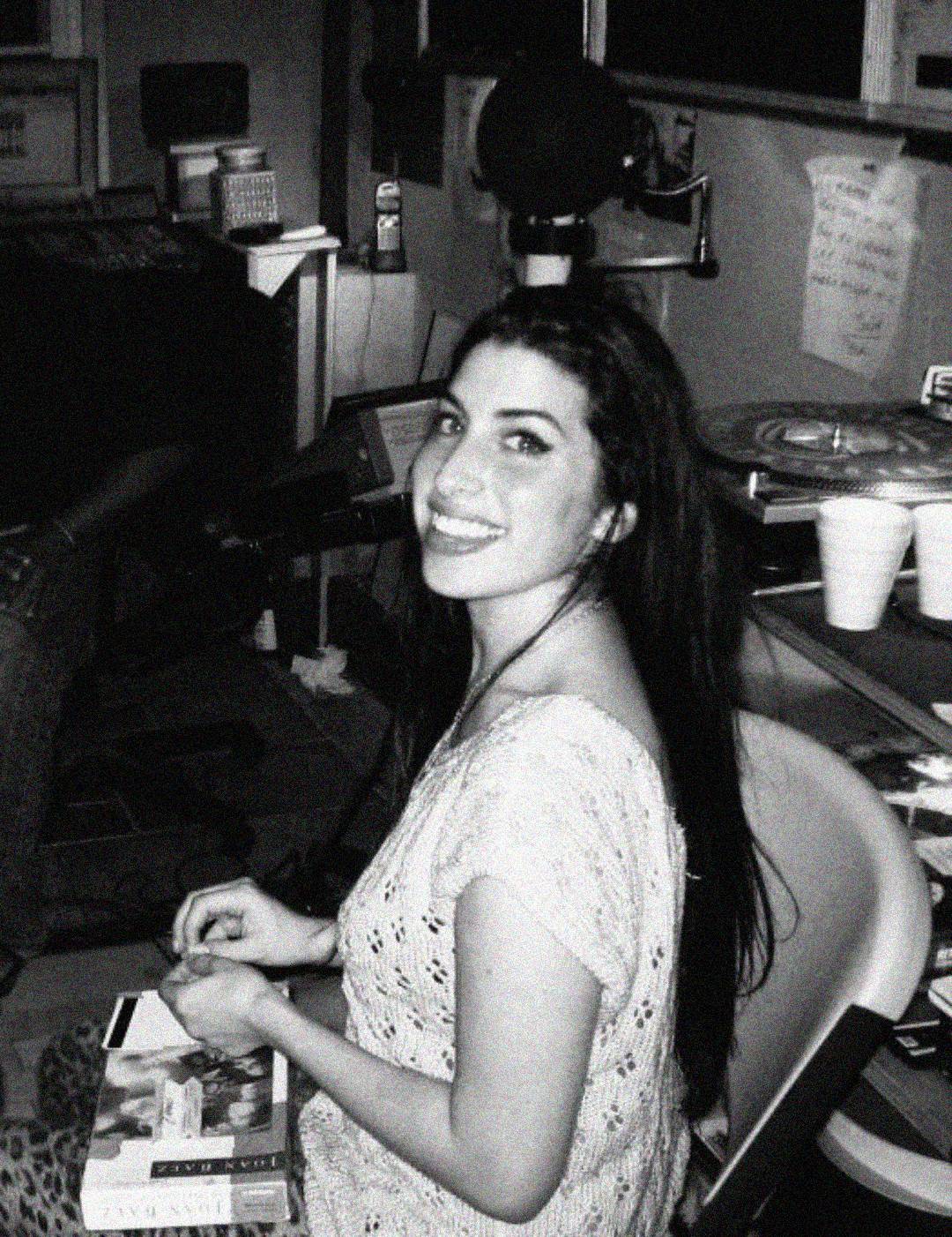 A young woman with long dark hair sits smiling at a desk in a cluttered room, holding a book. Two white cups and papers are on the desk. The photo is black and white and has a candid, vintage feel.