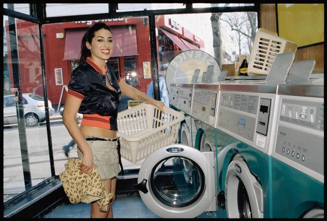 A smiling woman stands in a laundromat, holding a laundry basket and a leopard print bag. She is wearing a black cropped jacket and a khaki skirt, with washing machines and large windows behind her.