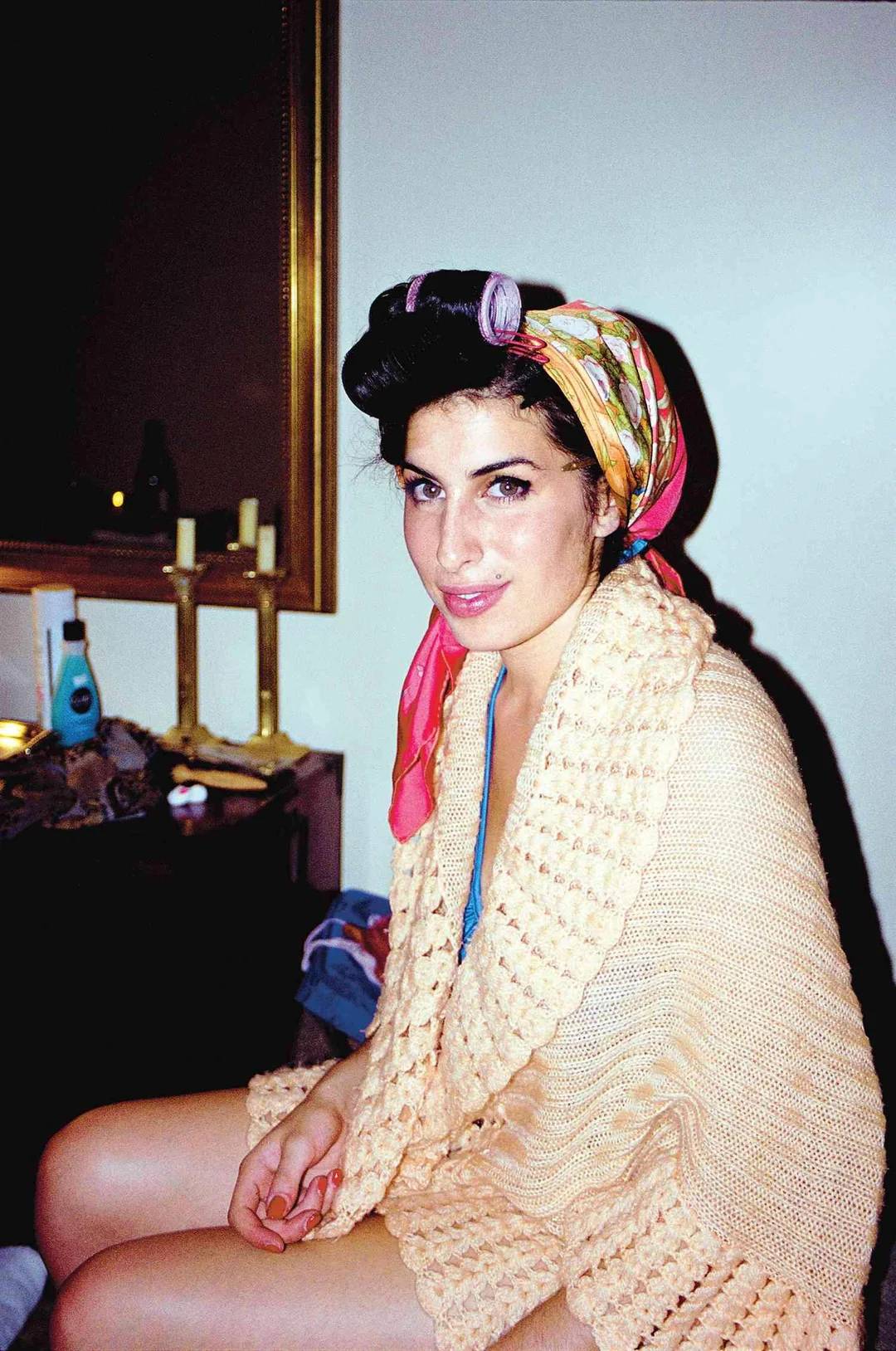 A woman with dark hair in curlers and a colorful scarf sits on a chair, wrapped in a beige knitted shawl. She looks at the camera and smiles softly. A mirror and candles are visible in the background.