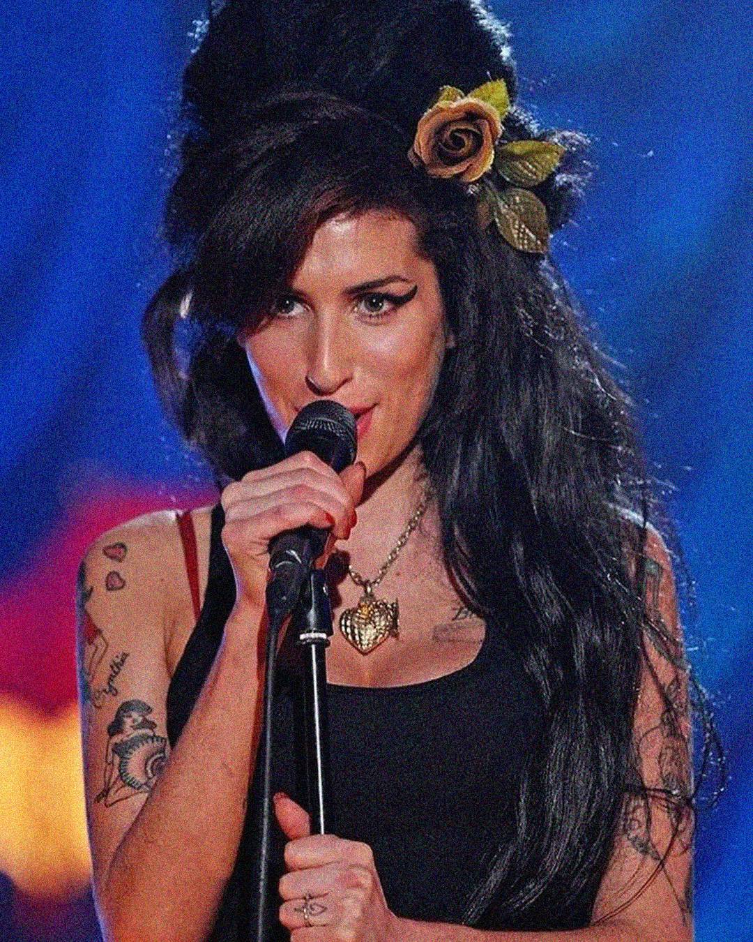 A woman with long dark hair styled in a beehive, wearing a black tank top and a rose hair accessory, sings into a microphone on stage. She has visible tattoos and is wearing a heart-shaped necklace.