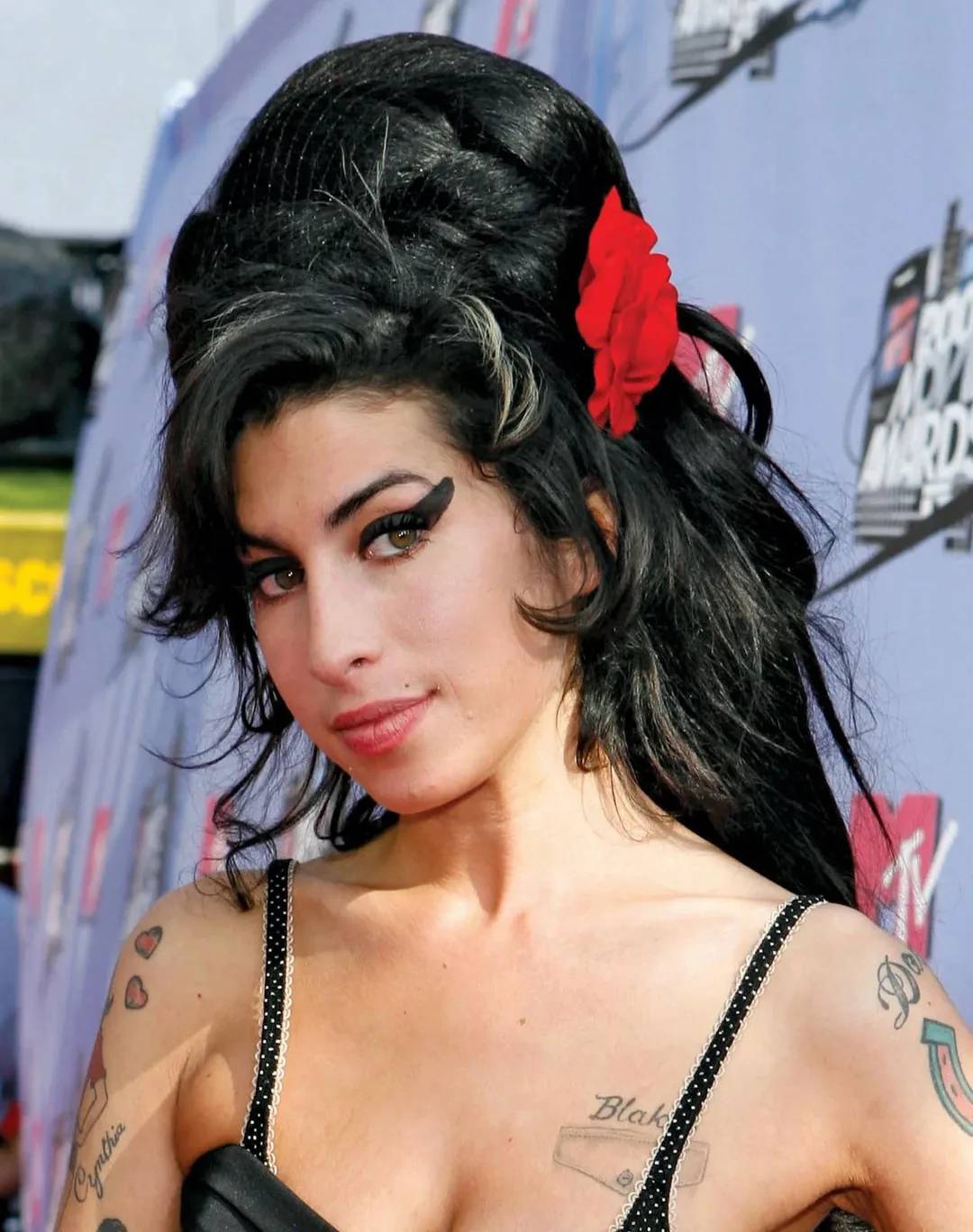 A woman with thick black hair in a beehive style, a red flower hair accessory, bold winged eyeliner, and tattoos on her arms stands in front of an MTV backdrop, wearing a black spaghetti-strap dress.