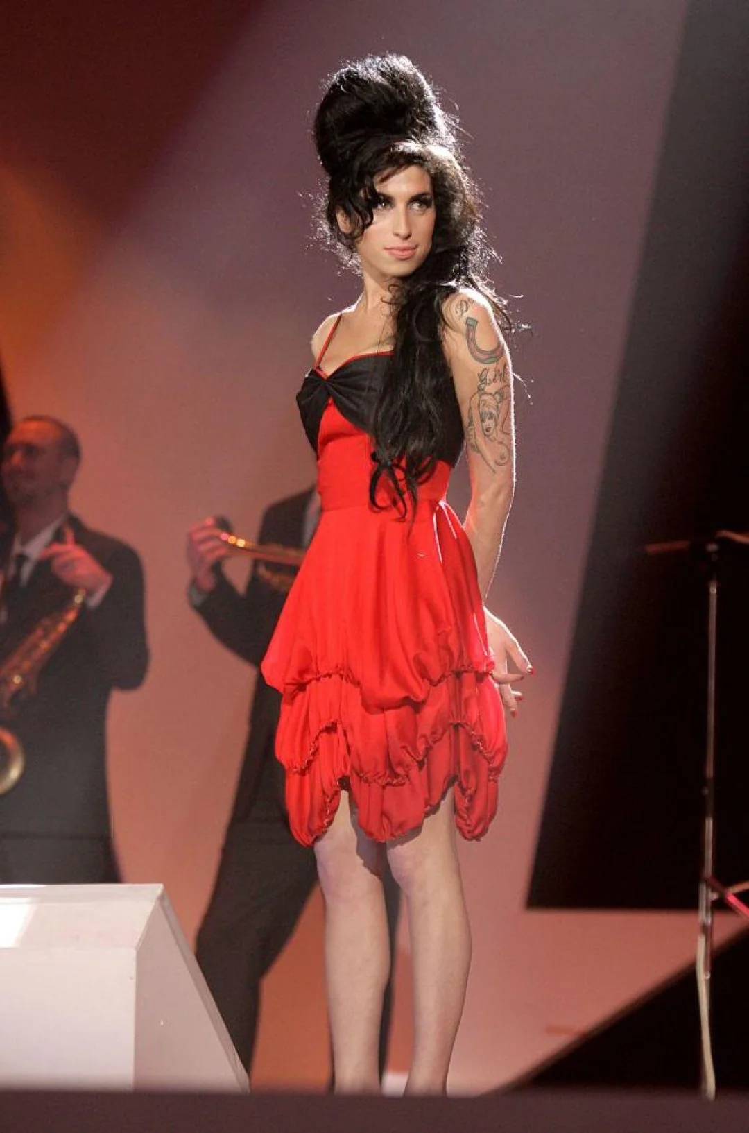A woman with long black hair in a beehive style wears a red and black dress, standing on stage. She has tattoos on her left arm, and musicians in suits play instruments in the background under stage lights.