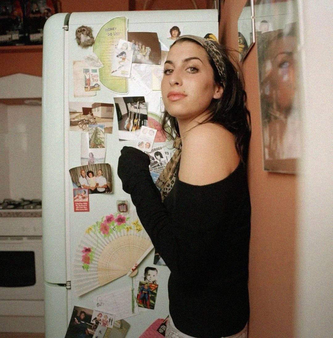 A woman with long dark hair and an off-the-shoulder black top stands in front of a refrigerator covered with photos, notes, and a decorative fan, in a cozy kitchen. She looks over her shoulder at the camera.