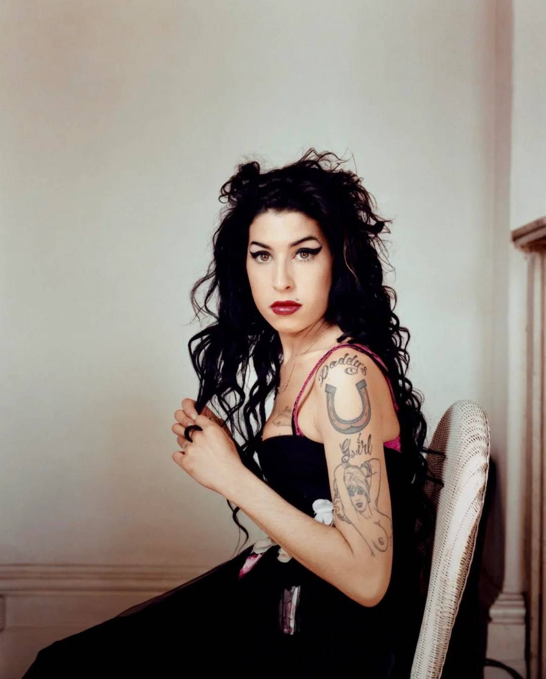A woman with long, curly black hair and visible tattoos on her arm sits sideways on a white chair, wearing a black strapless dress and looking toward the camera with a neutral expression.
