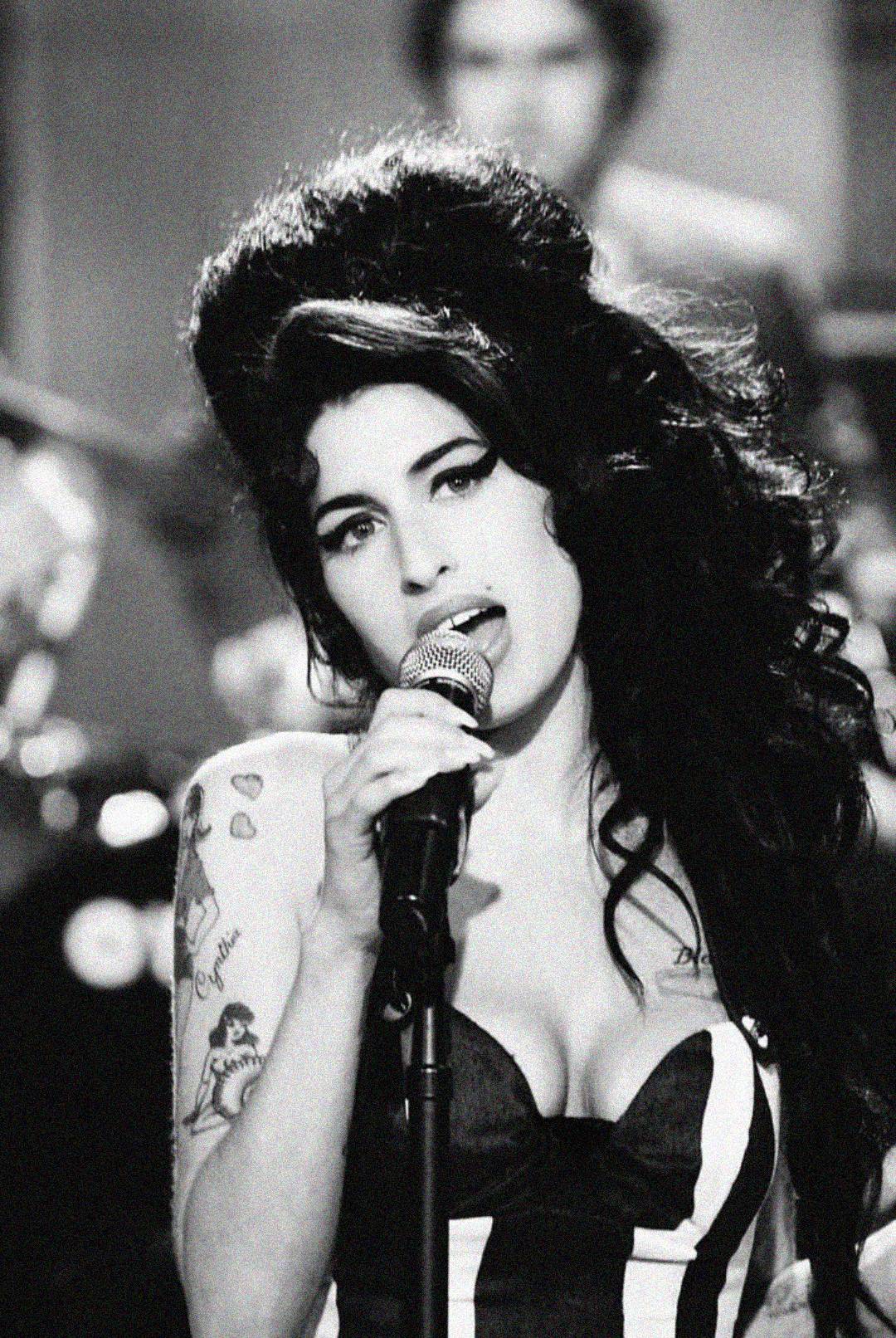Black and white photo of a woman with voluminous hair and tattoos, singing into a microphone. She is wearing a strapless dress with stripes and looking directly at the camera.