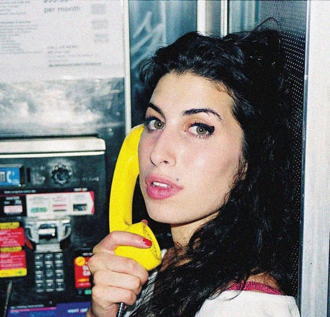 A woman with long dark hair holds a yellow payphone receiver to her ear, standing in front of a payphone. She has winged eyeliner and light pink lipstick, looking slightly to the side with a neutral expression.