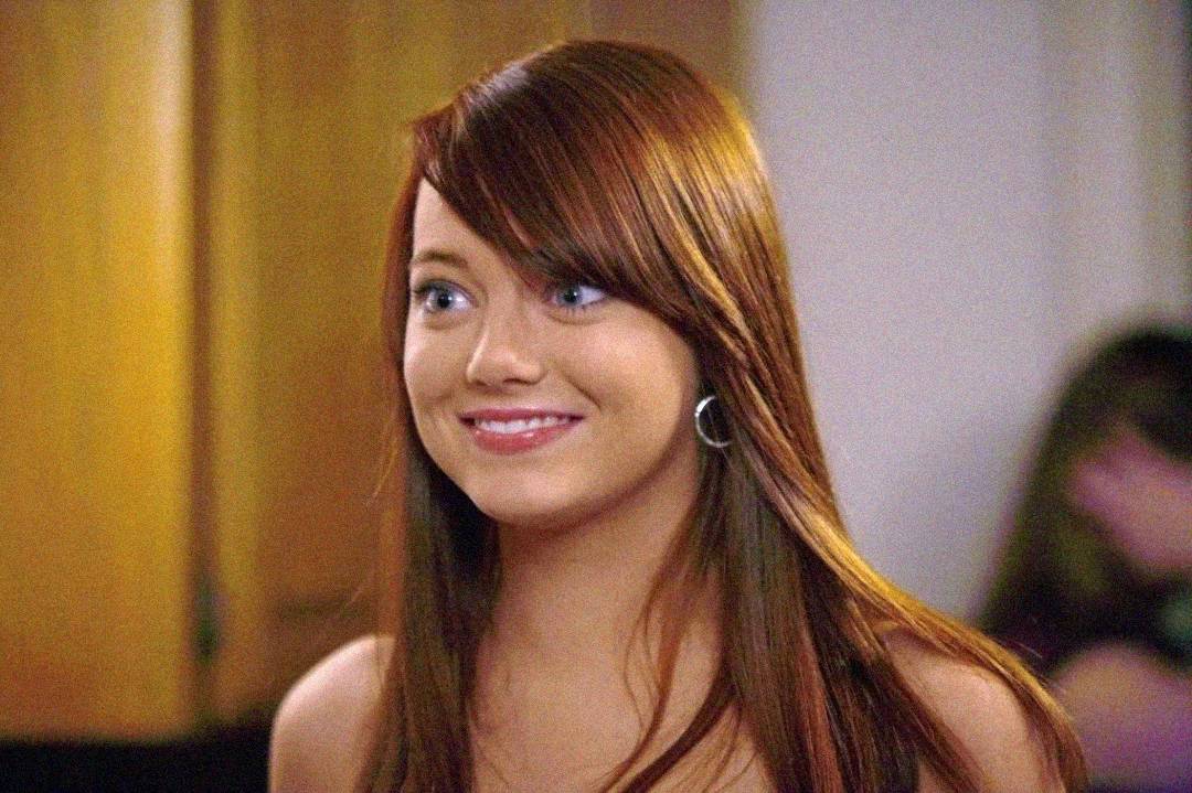 A young woman with straight, long auburn hair and blue eyes smiles slightly. She is wearing hoop earrings and standing indoors with a blurred background.