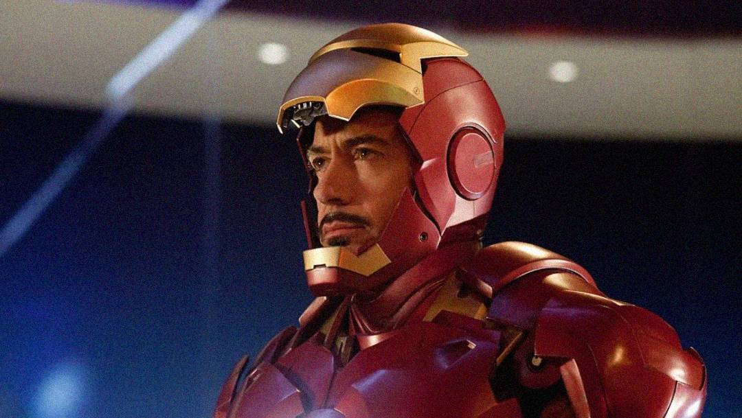 A man wearing a red and gold Iron Man suit with the helmet partially open, revealing his face. He has a serious expression and a neatly trimmed beard, standing in a futuristic, softly lit environment.