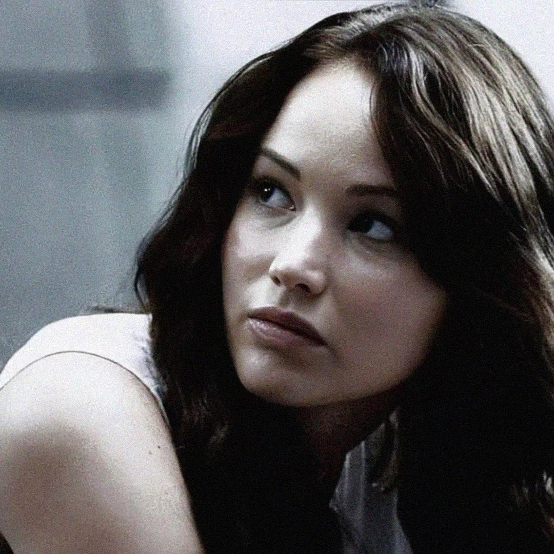 A young woman with long brown hair looks to her right with a serious, contemplative expression. The background is blurred and gray, emphasizing her focused face.