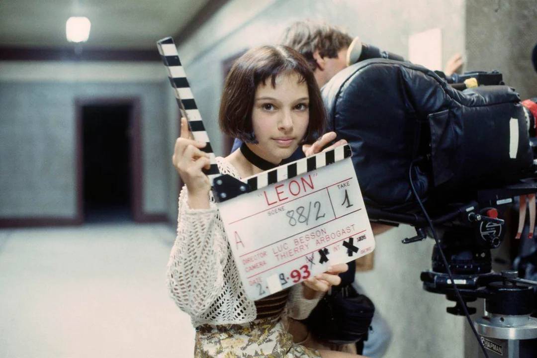 A young girl with short brown hair holds a film clapperboard labeled "LEON" in front of a camera on a movie set, with a blurred hallway in the background.