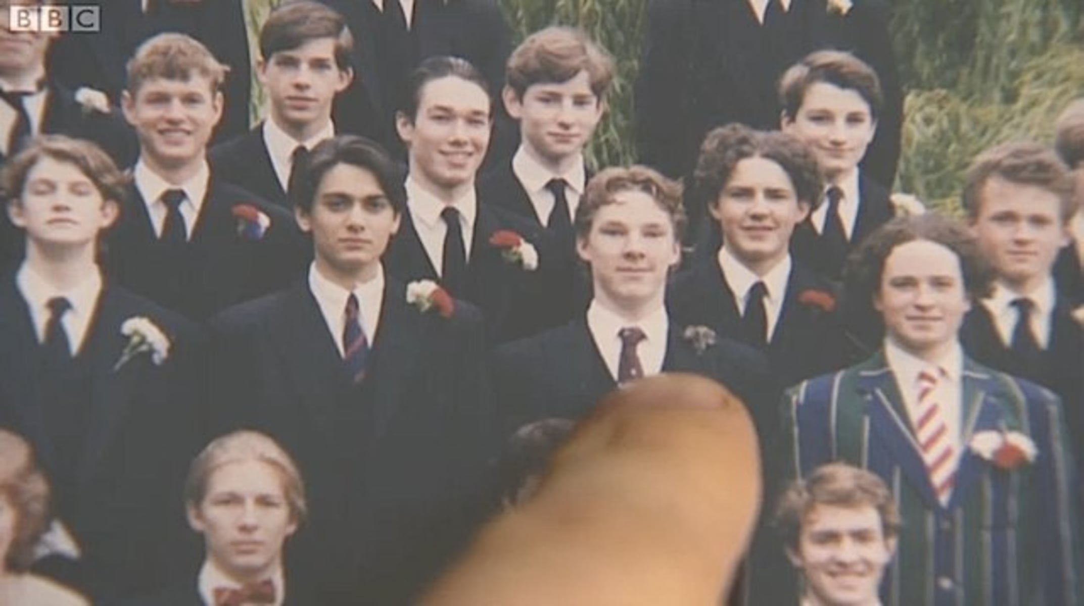 A finger points to a young man in a group of students dressed in formal suits and ties, some with boutonnieres, posing for a group photo outdoors. One student in the front wears a distinctive striped blazer.
