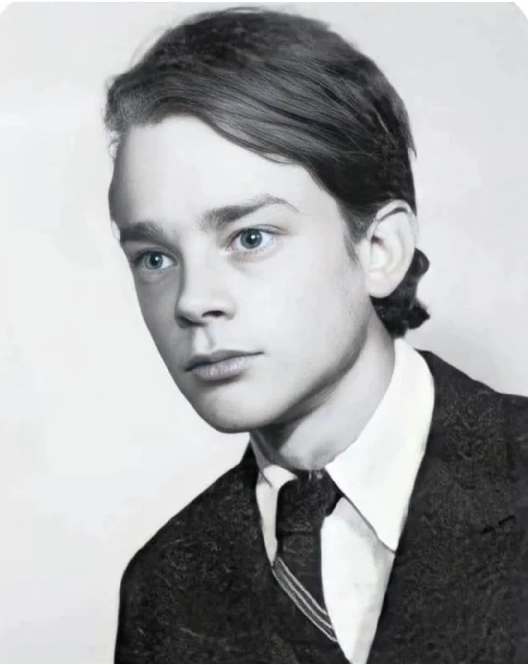 A black-and-white portrait of a young boy with light eyes and neatly combed dark hair, wearing a suit jacket, white collared shirt, and a striped tie, looking slightly to the side.