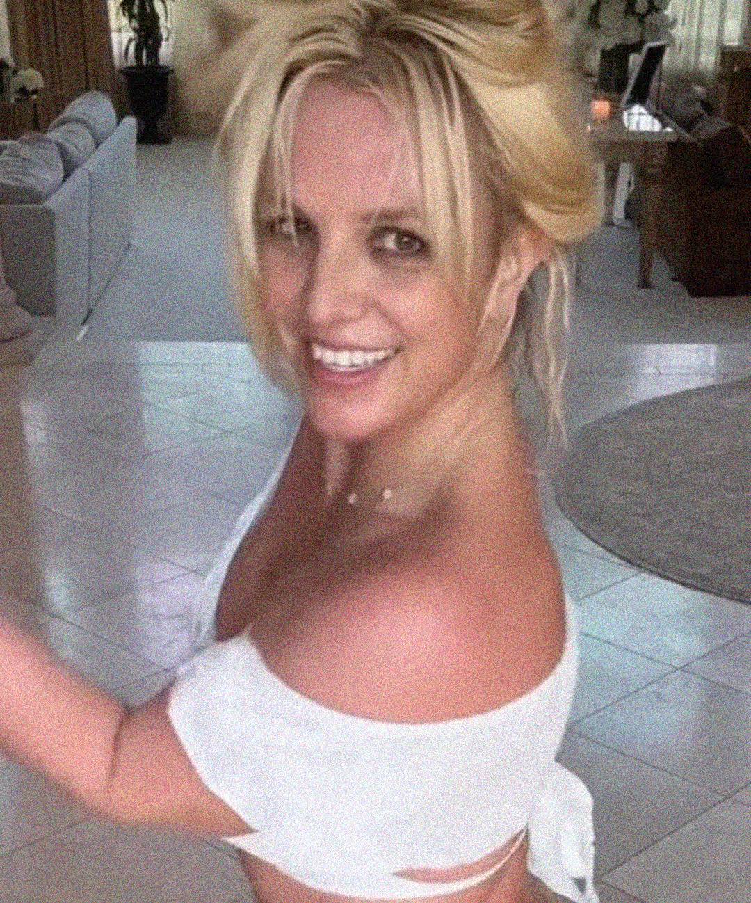 A woman with blonde hair in a messy updo smiles at the camera, wearing an off-the-shoulder white top, standing indoors on a tiled floor with a living room visible in the background.