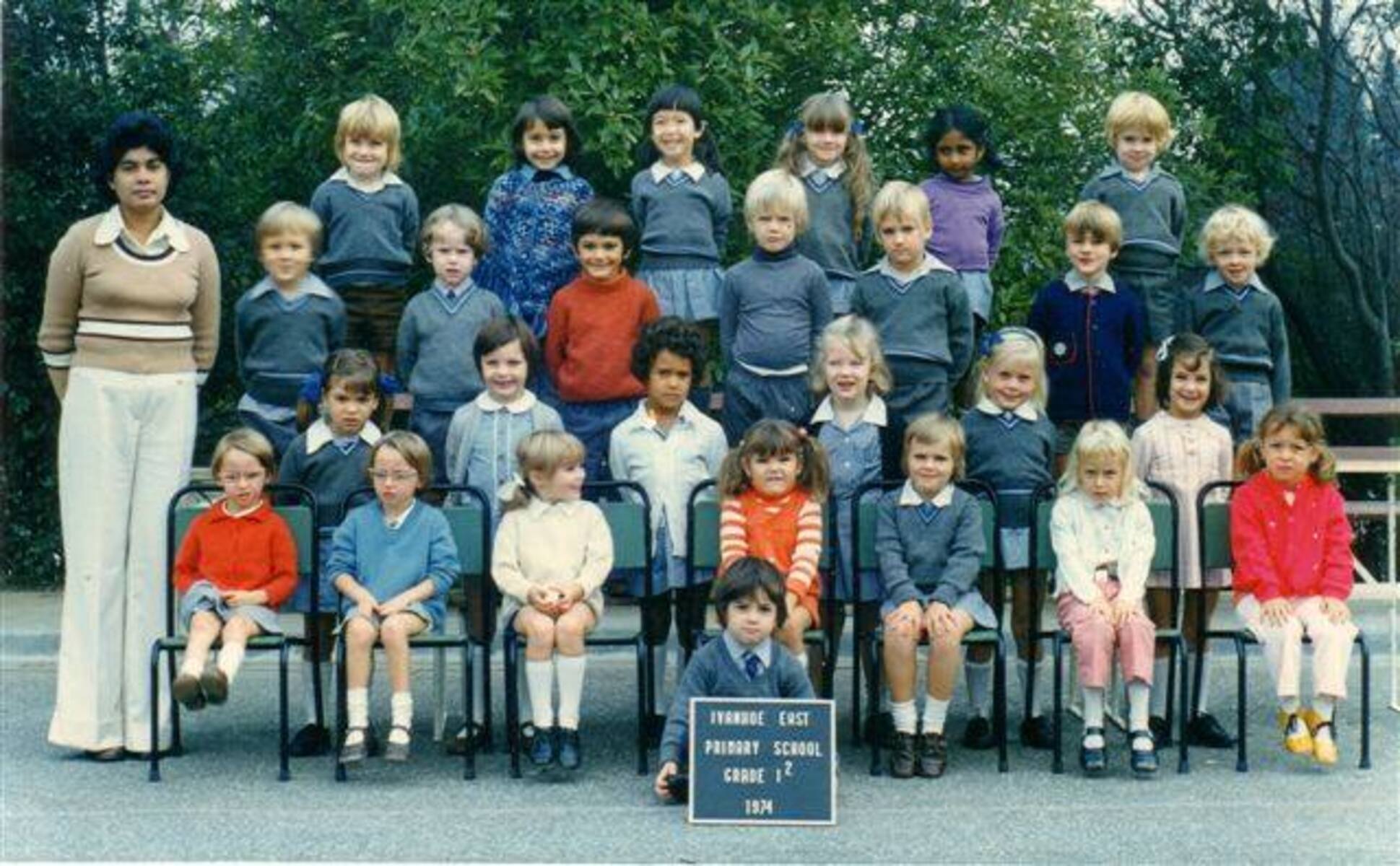A group of young children and a teacher pose for a class photo outdoors. The children sit and stand in rows, wearing school uniforms. A sign in front reads "Hyndale East Primary School Grade 1/2 1984.