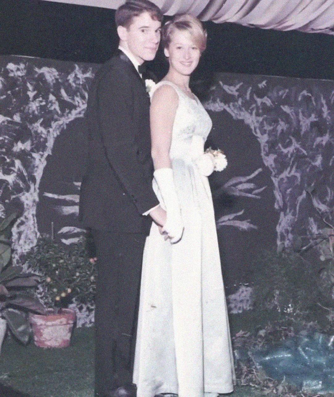 A young couple in formal attire poses together at what appears to be a prom or formal dance; the young man wears a dark suit and the young woman wears a light-colored gown with gloves. They are standing in front of a decorated backdrop.
