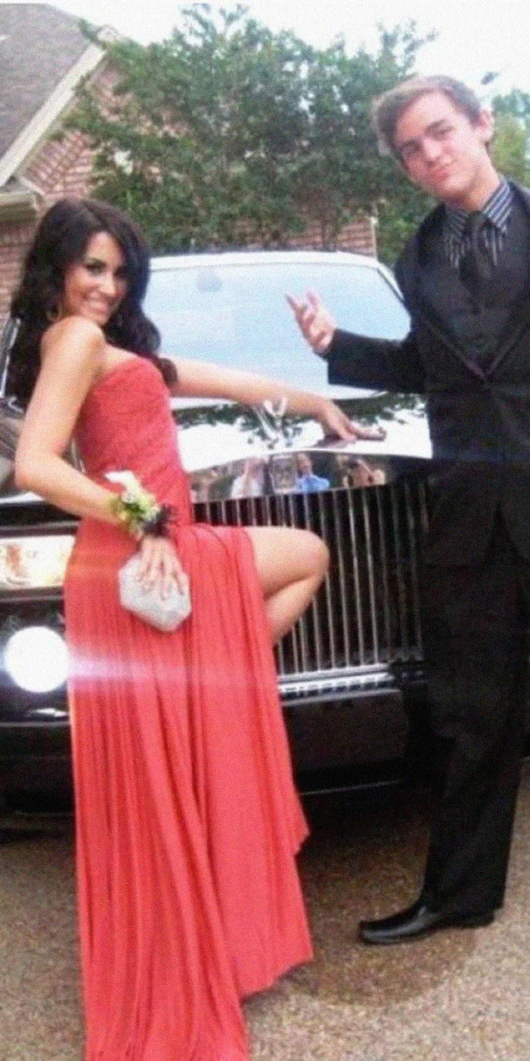 Two teenagers dressed in formal attire pose in front of a black luxury car. The young woman in a long red gown and corsage lifts her leg playfully, while the young man in a suit stands beside her, smiling and gesturing.
