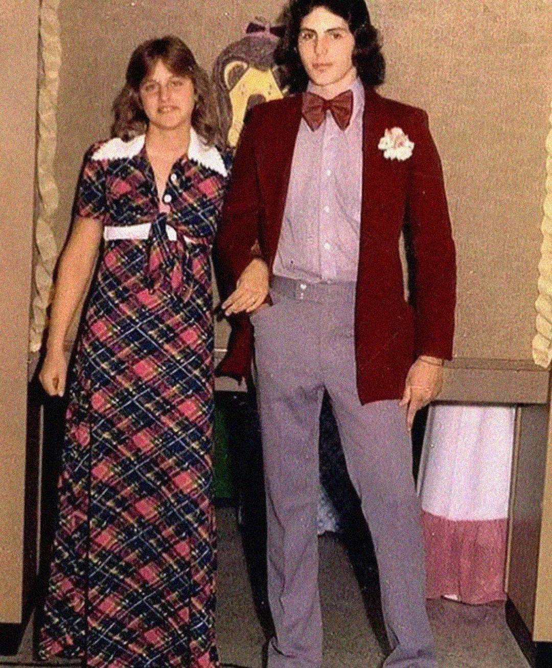 A young woman in a plaid dress stands next to a young man in a maroon jacket, bow tie, and gray pants. They are posing together indoors, likely at a formal event or dance.