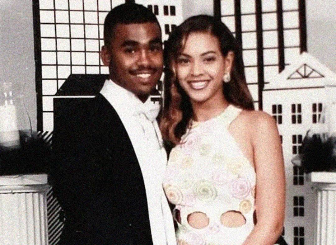 A young man in a black tuxedo and a young woman in a white dress with colorful swirls pose together, smiling, in front of a cityscape-themed backdrop.