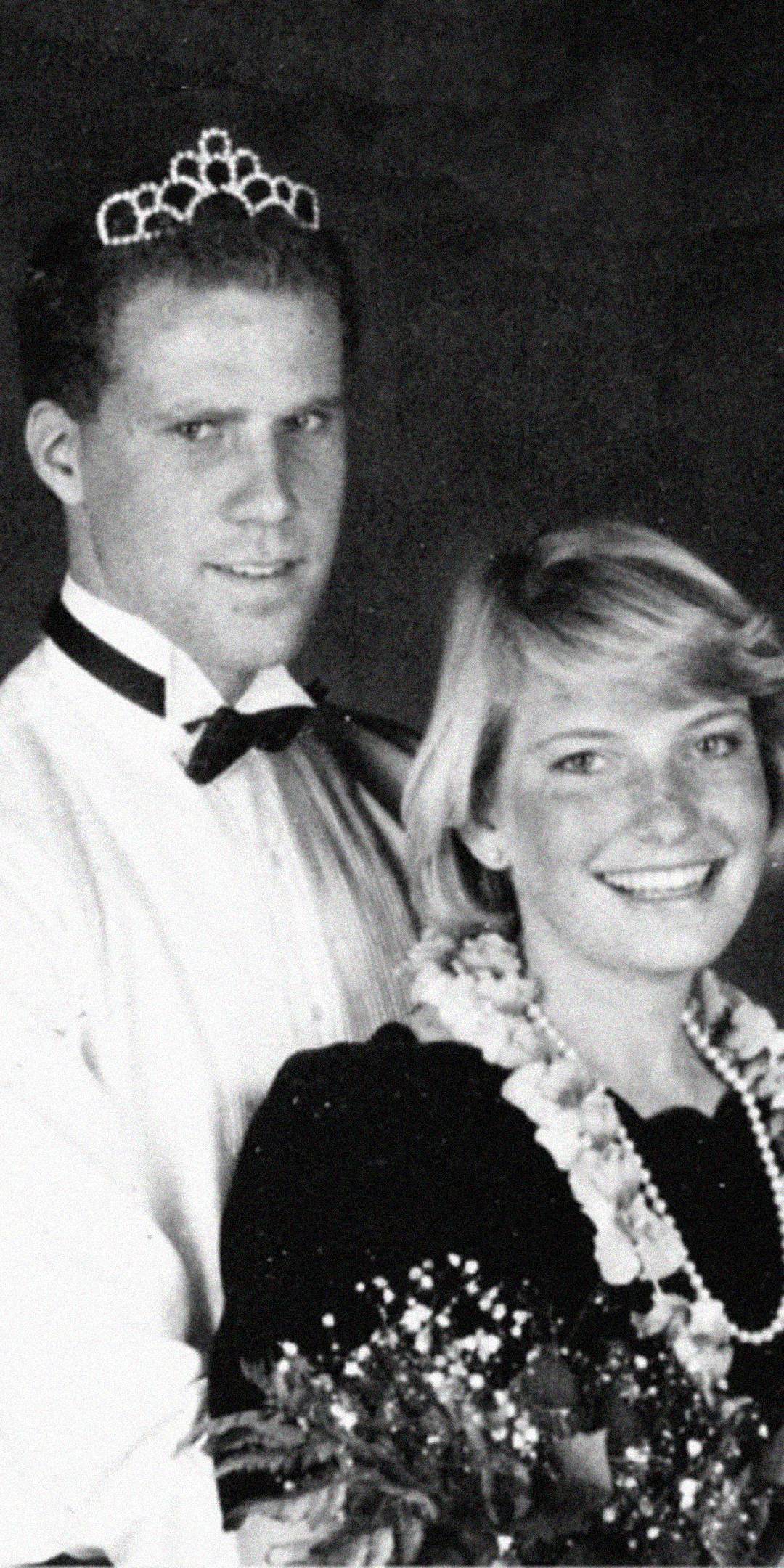 A black and white photo of a young man in a bow tie and a tiara standing behind a smiling young woman wearing a floral necklace and holding a bouquet, both dressed formally for what appears to be a special event.