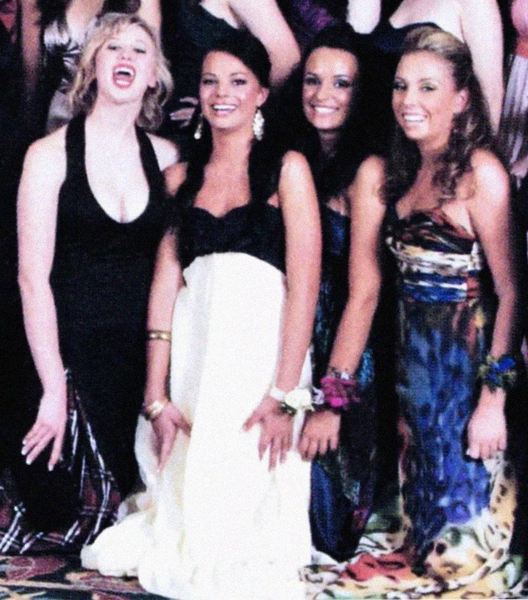 Four young women in colorful formal dresses posing closely together and smiling, likely at a prom or formal event. The background shows other people in similar attire.