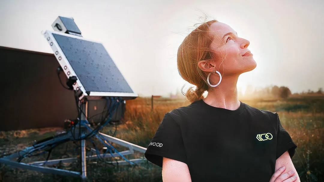 A woman with light hair in a ponytail, wearing a black t-shirt and large hoop earrings, stands outdoors at sunset, smiling and looking up. Behind her is a large technological device on a tripod in a grassy field.