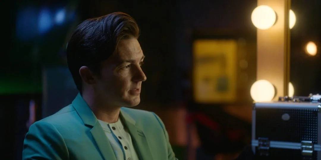 A man in a mint green blazer sits in front of an illuminated mirror, looking thoughtful. His hair is neatly styled and he wears a white shirt. The background is softly lit with yellow and green tones.