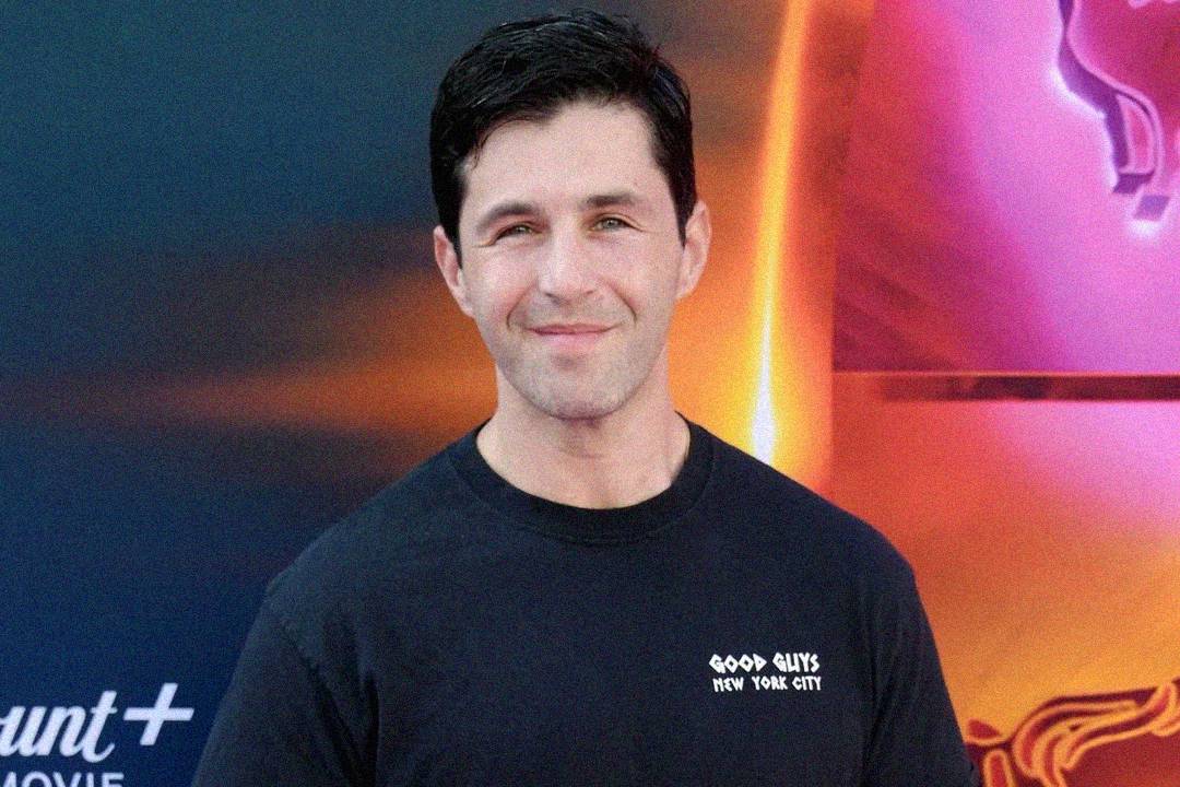 A man with short dark hair smiles at the camera. He is wearing a black T-shirt that reads "GOOD GUYS NEW YORK CITY" and is standing in front of a colorful, abstract background.