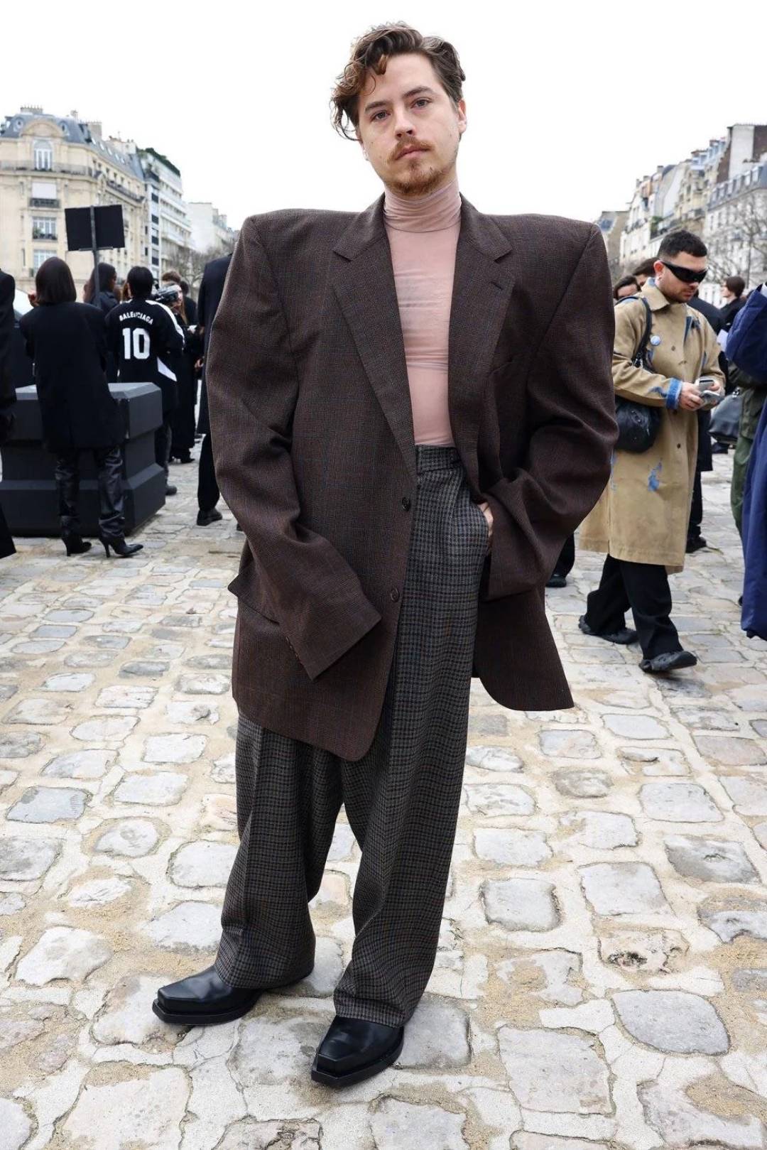 A person wearing a large brown blazer with exaggerated padded shoulders, a light pink turtleneck, and wide plaid trousers stands on a cobblestone street, surrounded by people in urban outdoor setting.
