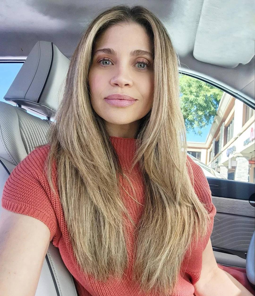A woman with long, straight blonde hair and light eyes sits in a car, wearing a short-sleeved rust-colored top. Sunlight enters through the window, and buildings and a tree are visible outside.
