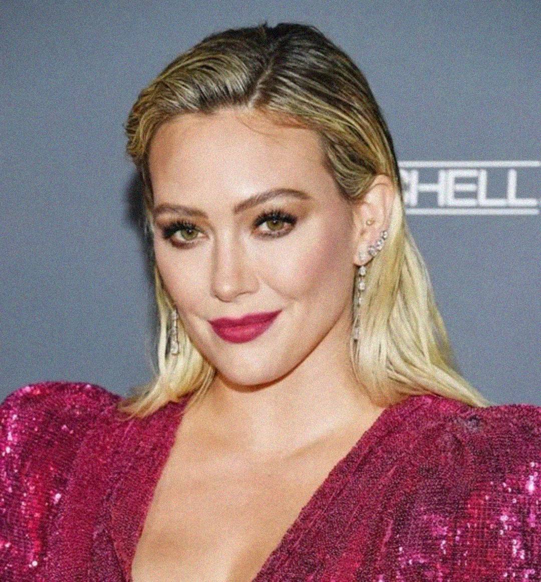 A woman with blonde hair in loose waves wears a sparkly, deep pink outfit and statement earrings, smiling slightly against a gray background at an event.
