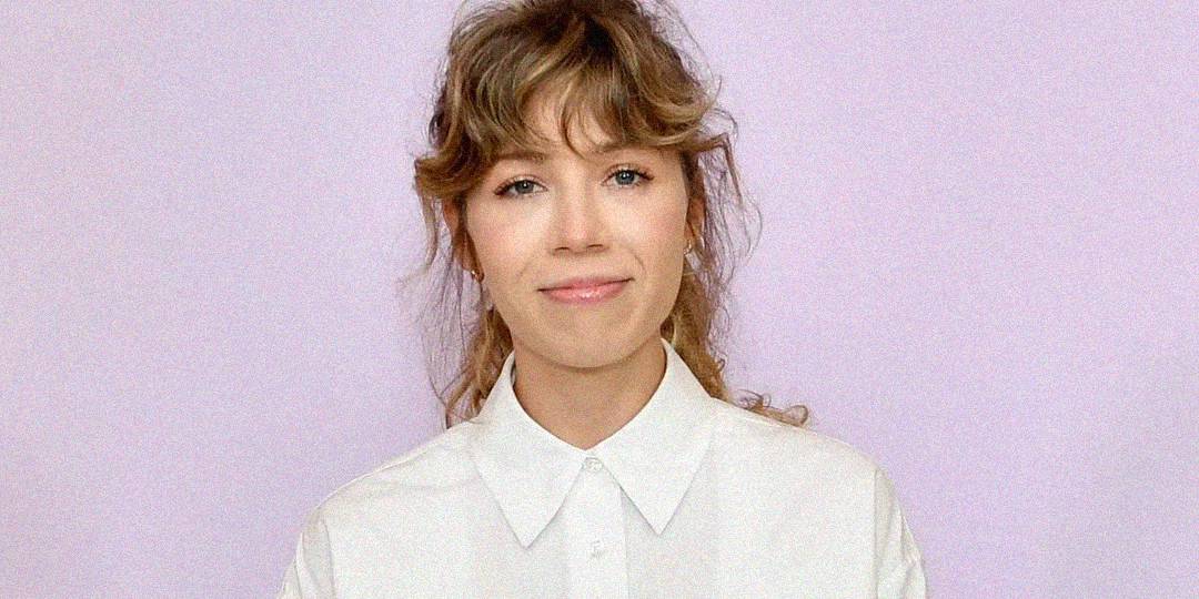 A young woman with wavy light brown hair and bangs smiles softly. She is wearing a white collared shirt and stands in front of a plain light purple background.